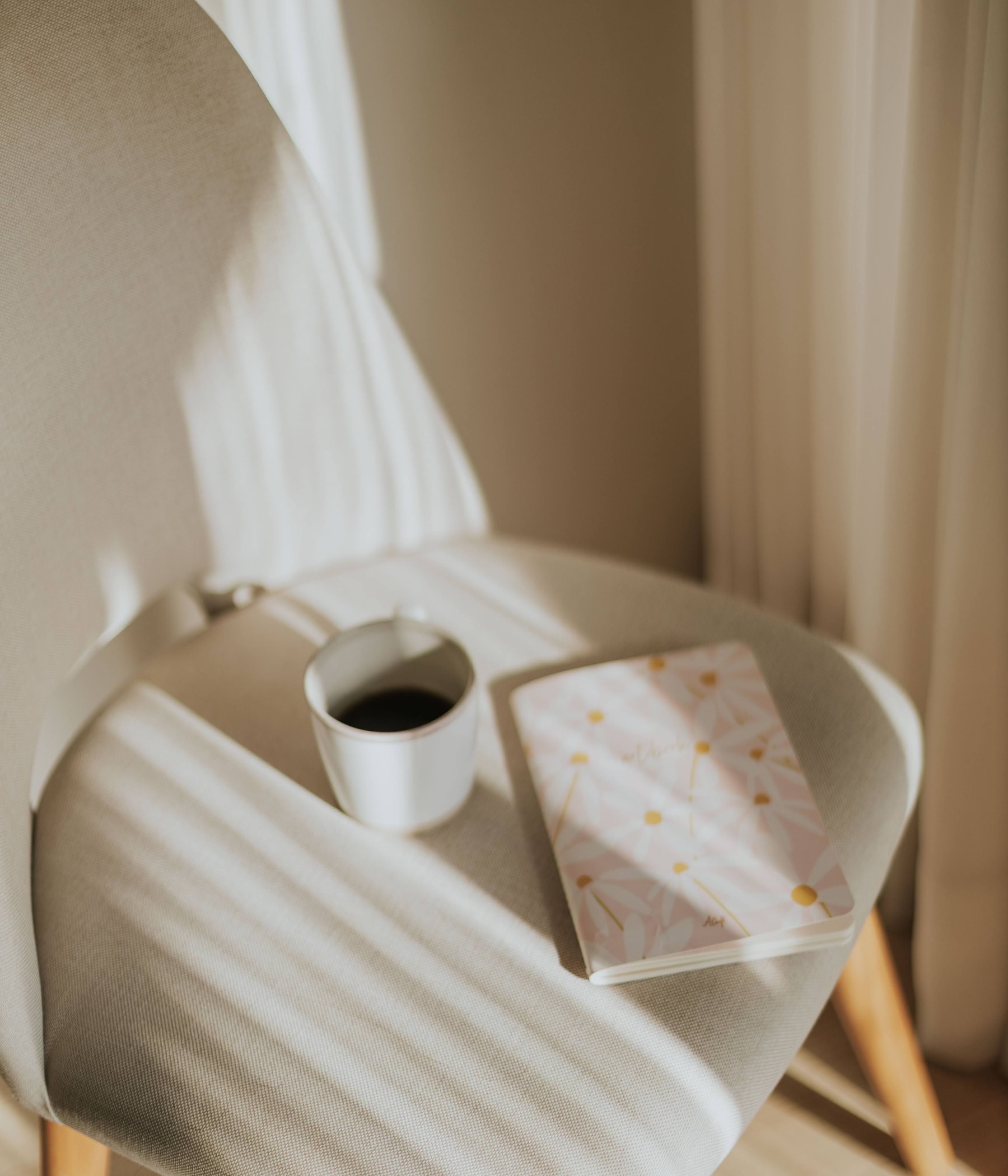 A cup of coffee and a notebook on a pale chair striped with sunlight.