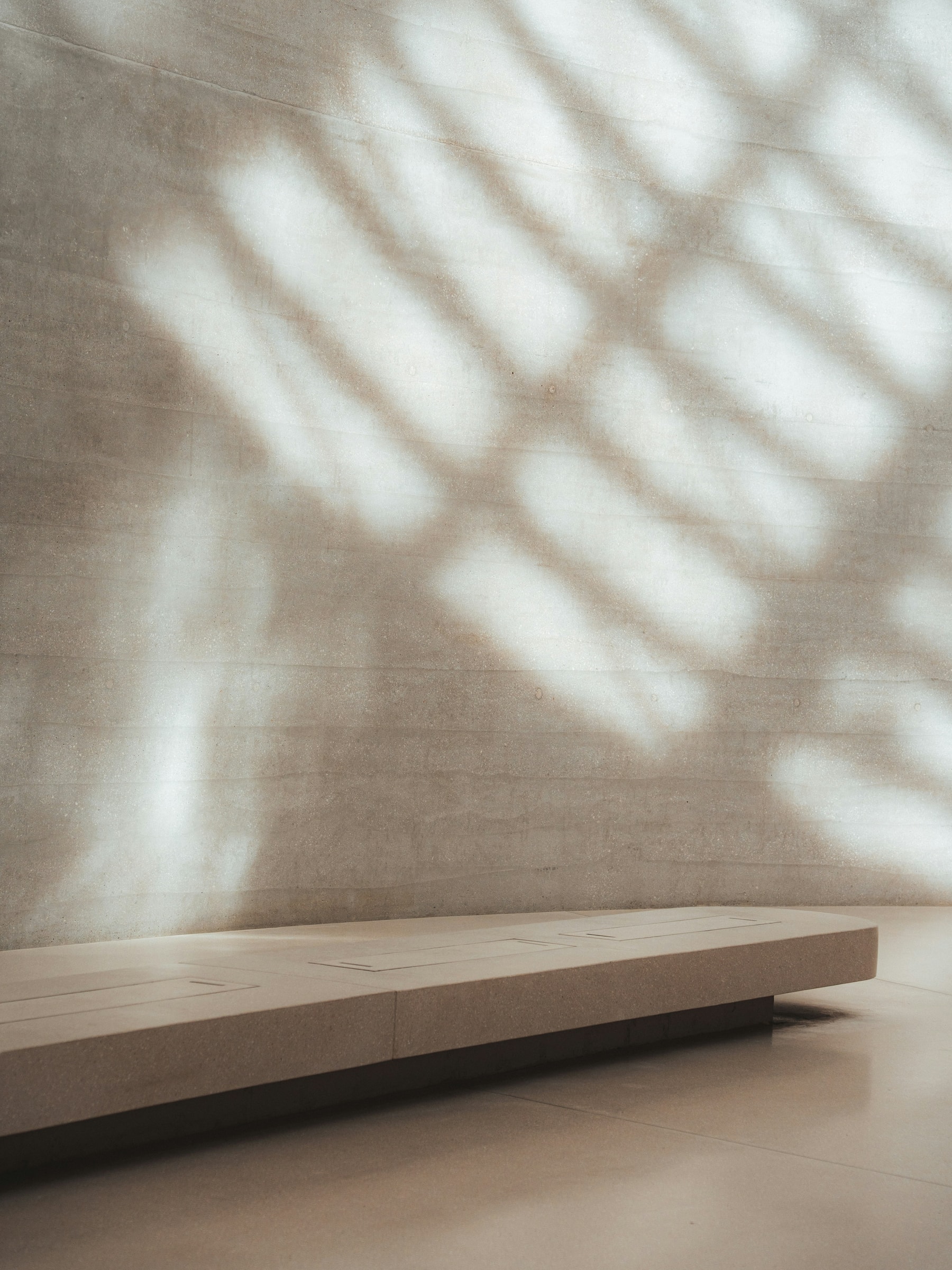 A long bench beneath a textured stone wall patterned with soft sunlight.