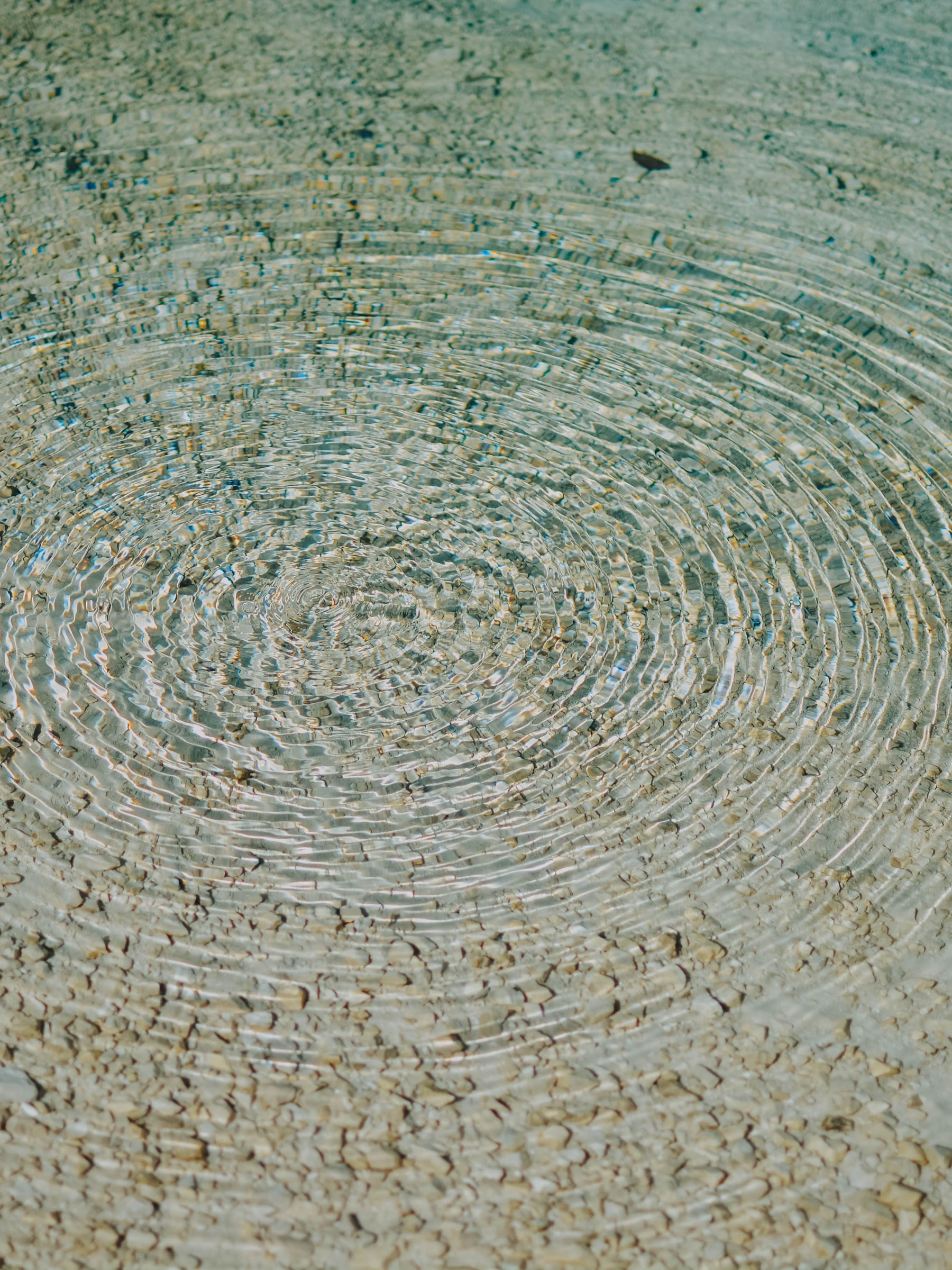 Concentric ripples spreading over pebbles in clear shallow water.